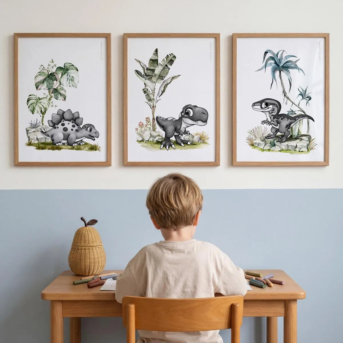 Trio of hand-drawn dinosaur nursery prints—Stegosaurus, T-Rex, and Velociraptor—in oak frames, styled above a child's wooden art desk in a modern blue and white playroom.