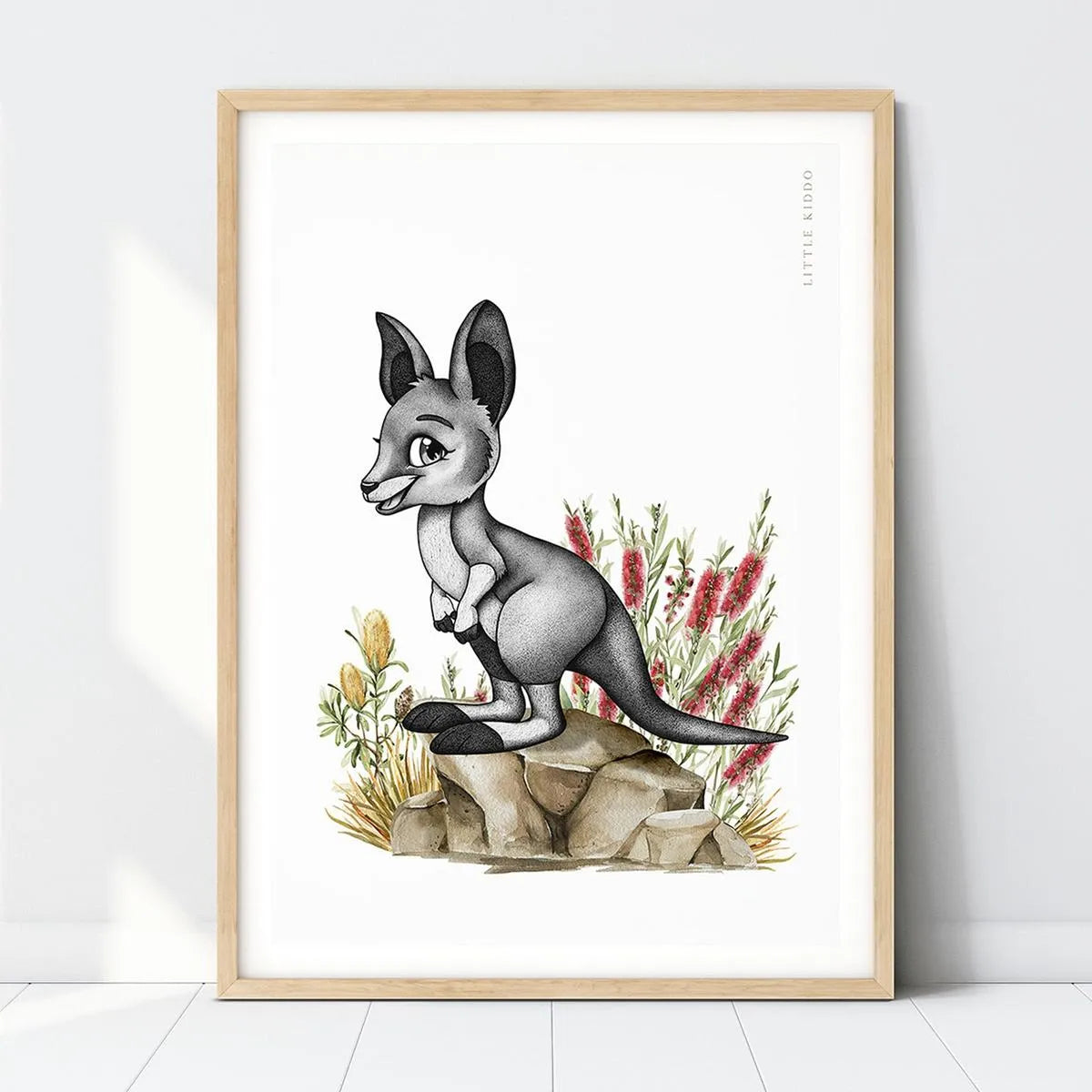 Hand-drawn baby kangaroo nursery art print standing on rocks among red bottle brush flowers in a light wood frame.