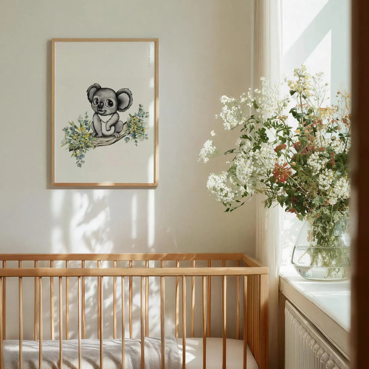 Sophisticated nursery wall art featuring a hand-drawn koala on a branch with yellow watercolor wattle flowers. The framed print is styled above a light timber baby crib in a sun-drenched room with a vase of white wildflowers in the foreground.