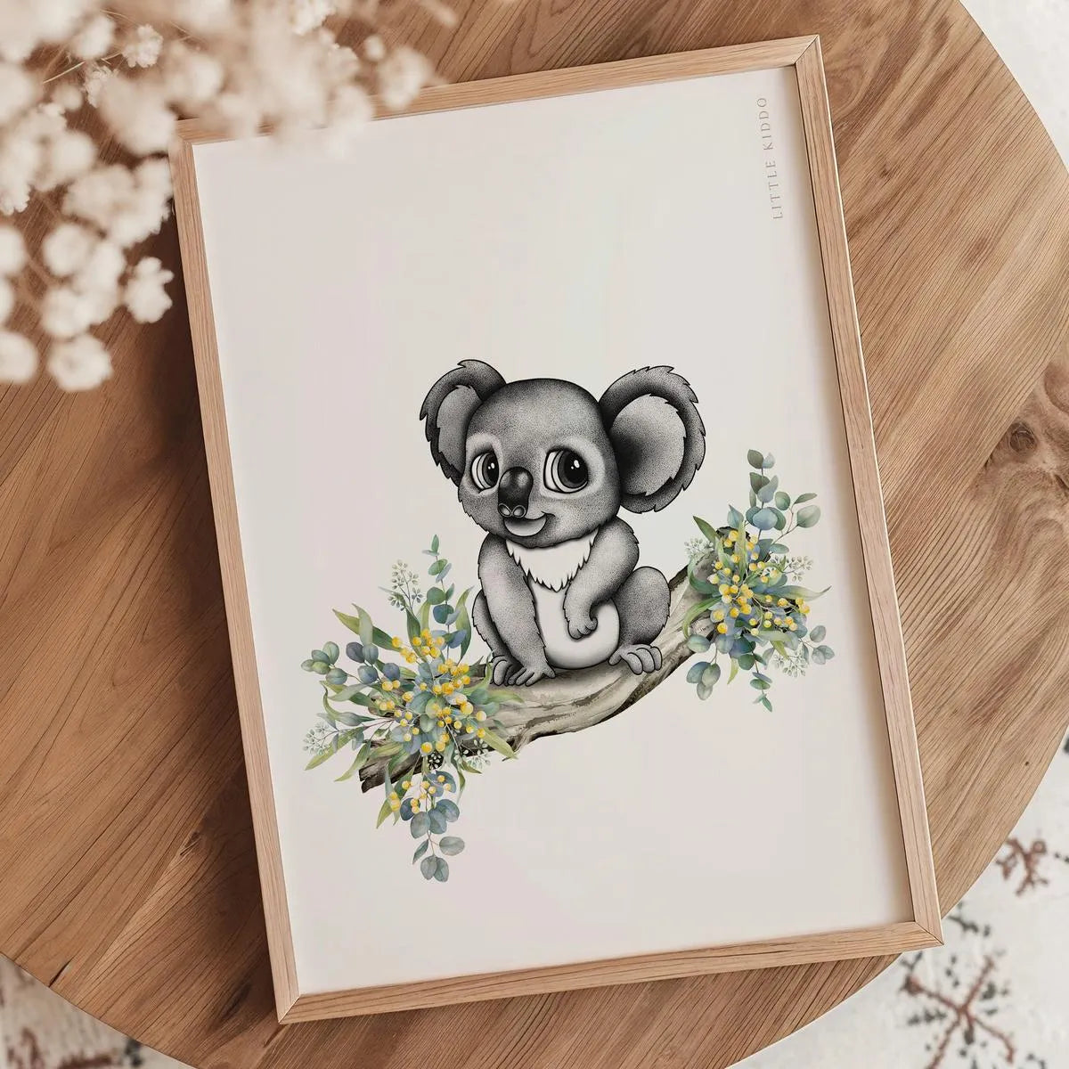 Minimalist koala illustration nursery print in an oak frame styled on a wooden tabletop with white dried botanical accents.