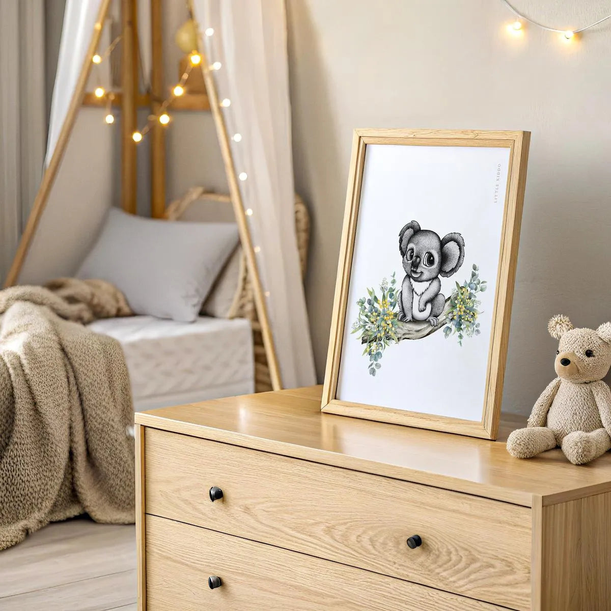 Sophisticated koala nursery print in a timber frame, styled on a wooden dresser with a teddy bear and warm fairy lights. Hand-drawn with black and white dots and watercolor flowers.