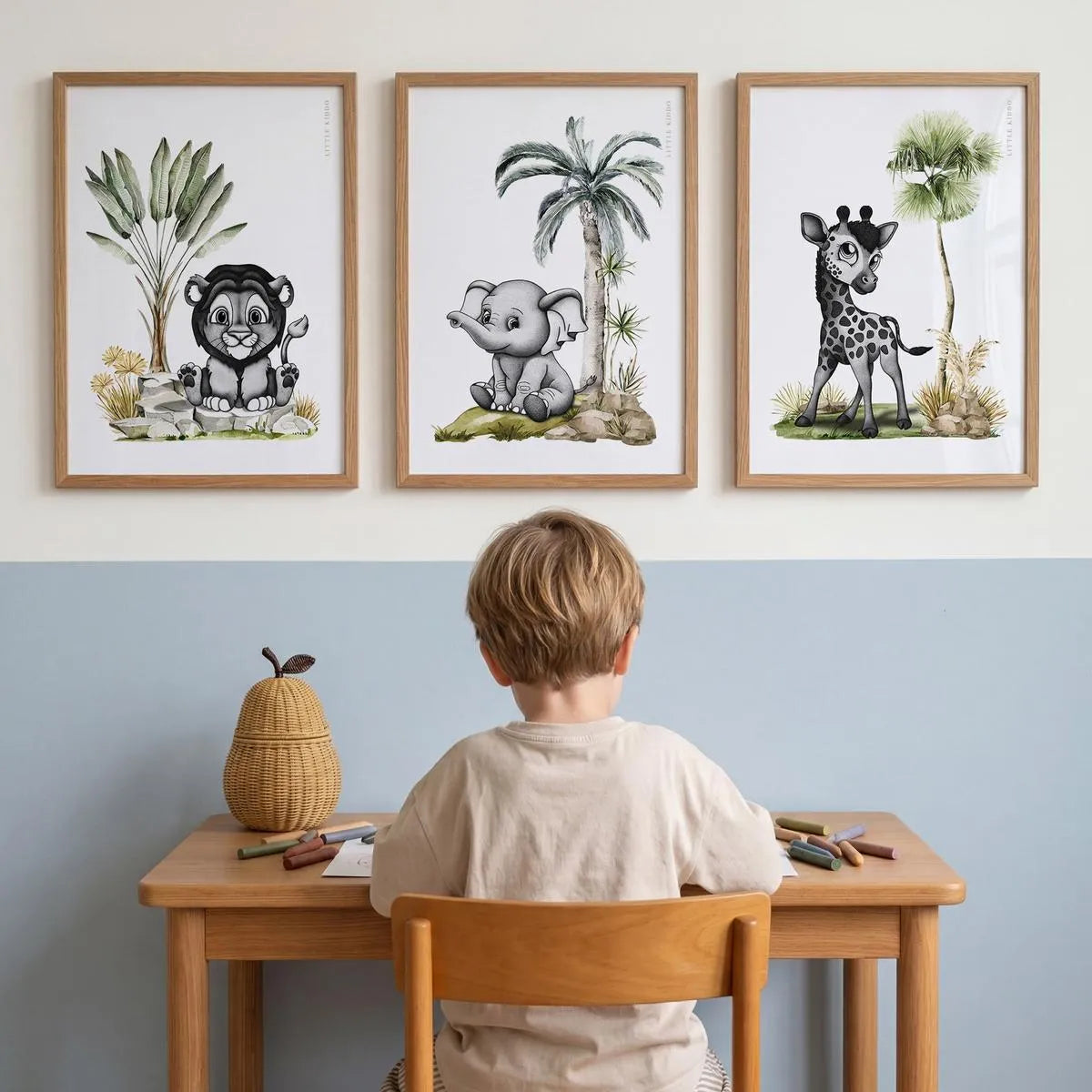 A young boy sitting at a desk below a gallery wall of three framed safari animal prints. The sophisticated nursery decor includes a hand-drawn lion, elephant, and giraffe, created with intricate stippling and soft watercolor greenery.