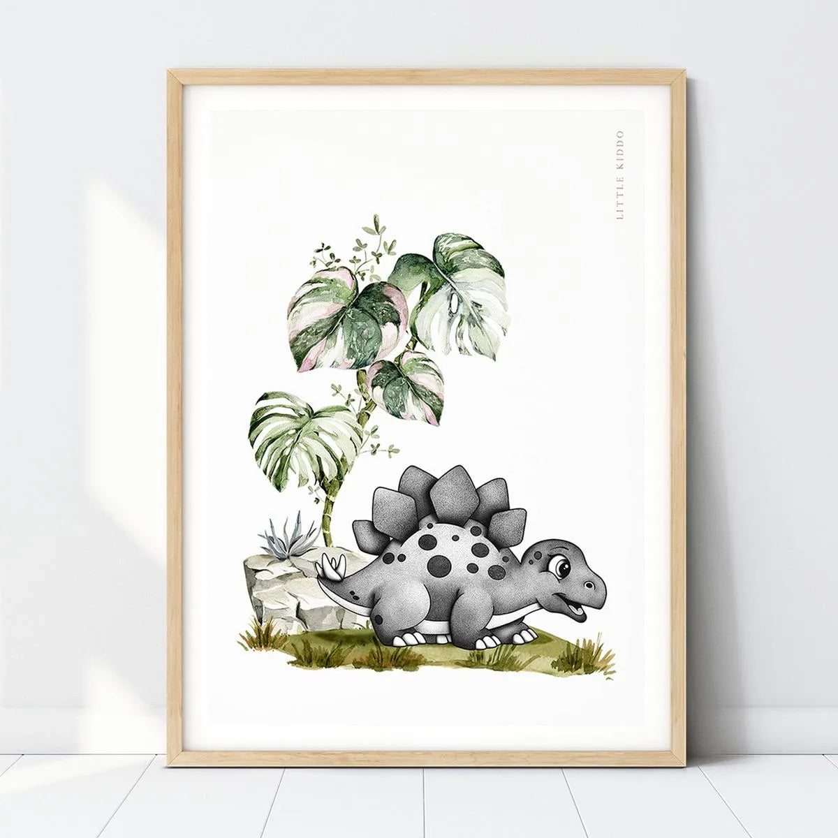 Hand-drawn baby Stegosaurus nursery art print under a Monstera leaf in a light wood frame, prehistoric dinosaur wall decor.