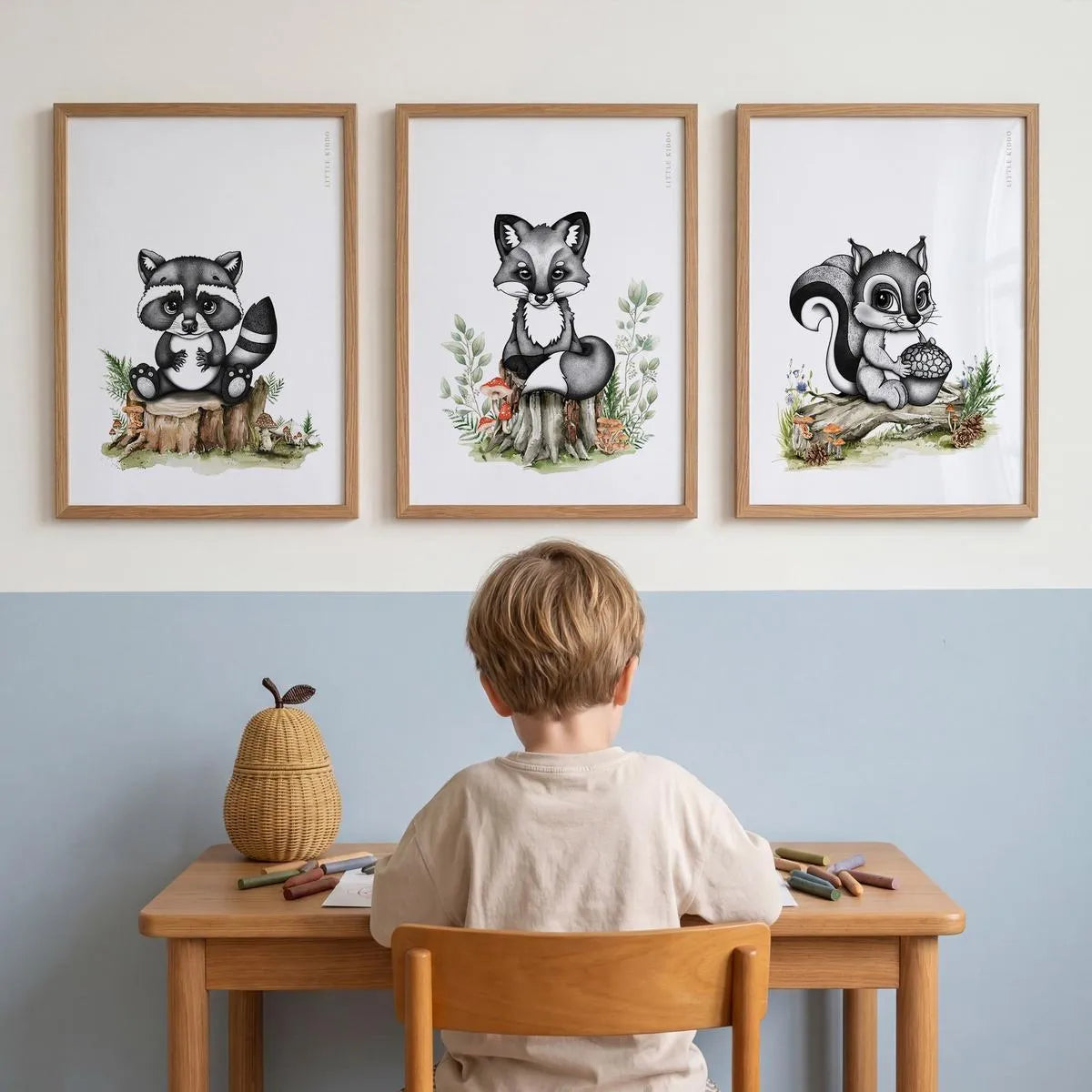 Set of 3 woodland nursery prints—raccoon, fox, and squirrel—in oak frames, styled above a child's wooden art desk in a modern blue and white playroom.