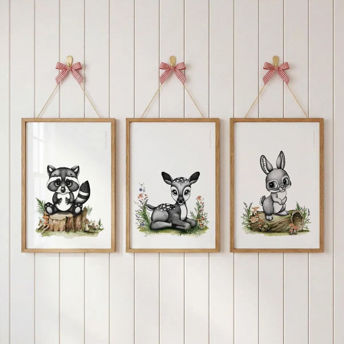 Set of 3 woodland nursery prints—raccoon, deer, and bunny-rabbit—hanging with pink ribbons on a white wood-paneled wall.