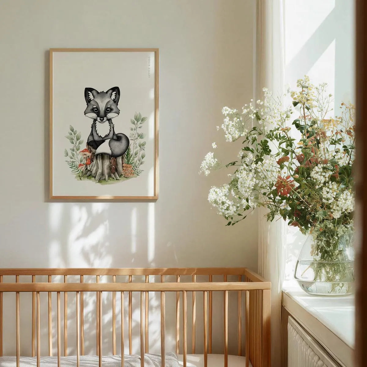 Hand-drawn baby fox nursery art print in a light wood frame hanging above a wooden crib, styled with fresh white flowers in a sunlit nursery.