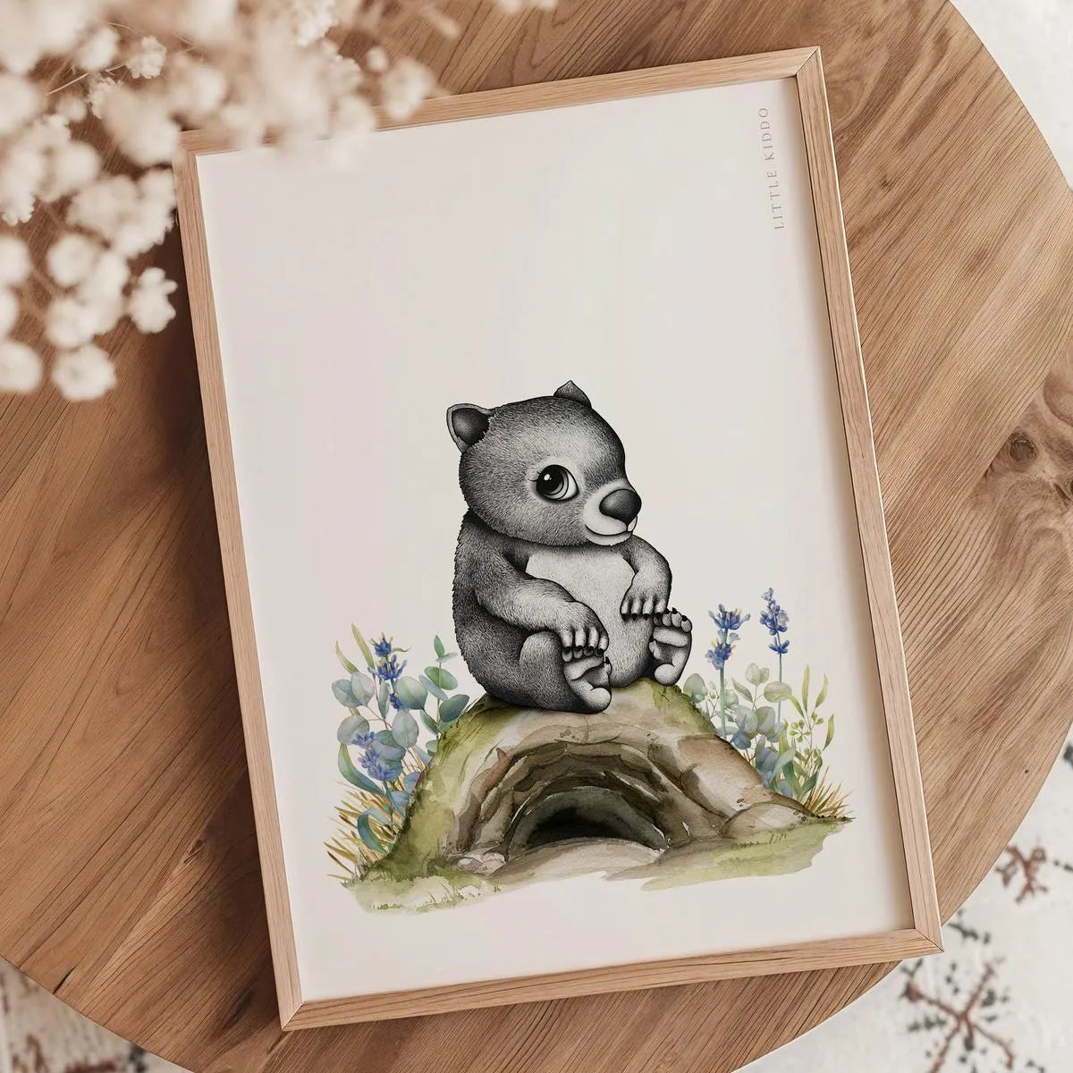 Minimalist wombat illustration nursery print in an oak frame styled on a wooden tabletop with white dried flowers and botanical accents.
