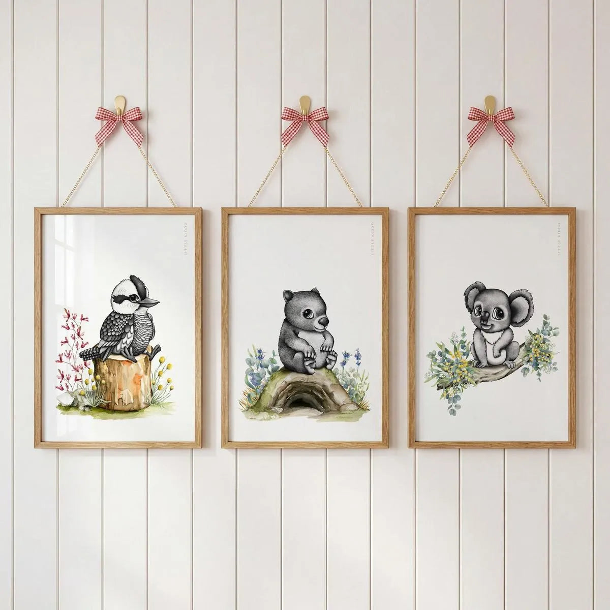 Gallery wall set of 3 Australian nursery prints—kookaburra, wombat, and koala—hanging with pink ribbons on a white paneled wall.