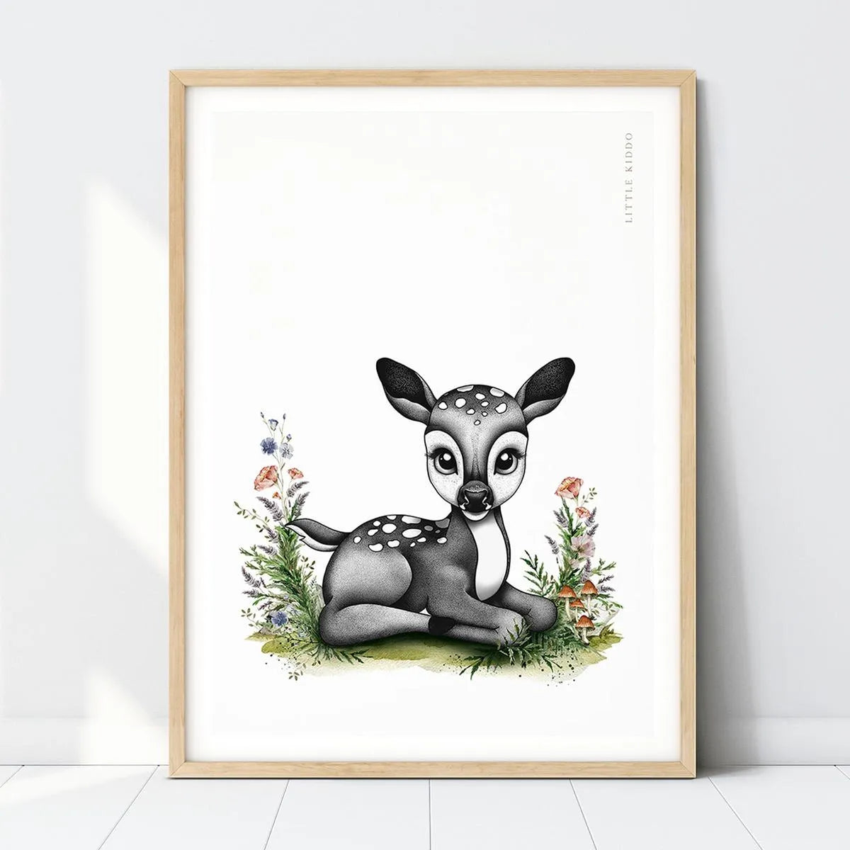 Hand-drawn baby fawn nursery art print resting in a meadow with wildflowers in a light wood frame, woodland deer wall decor.