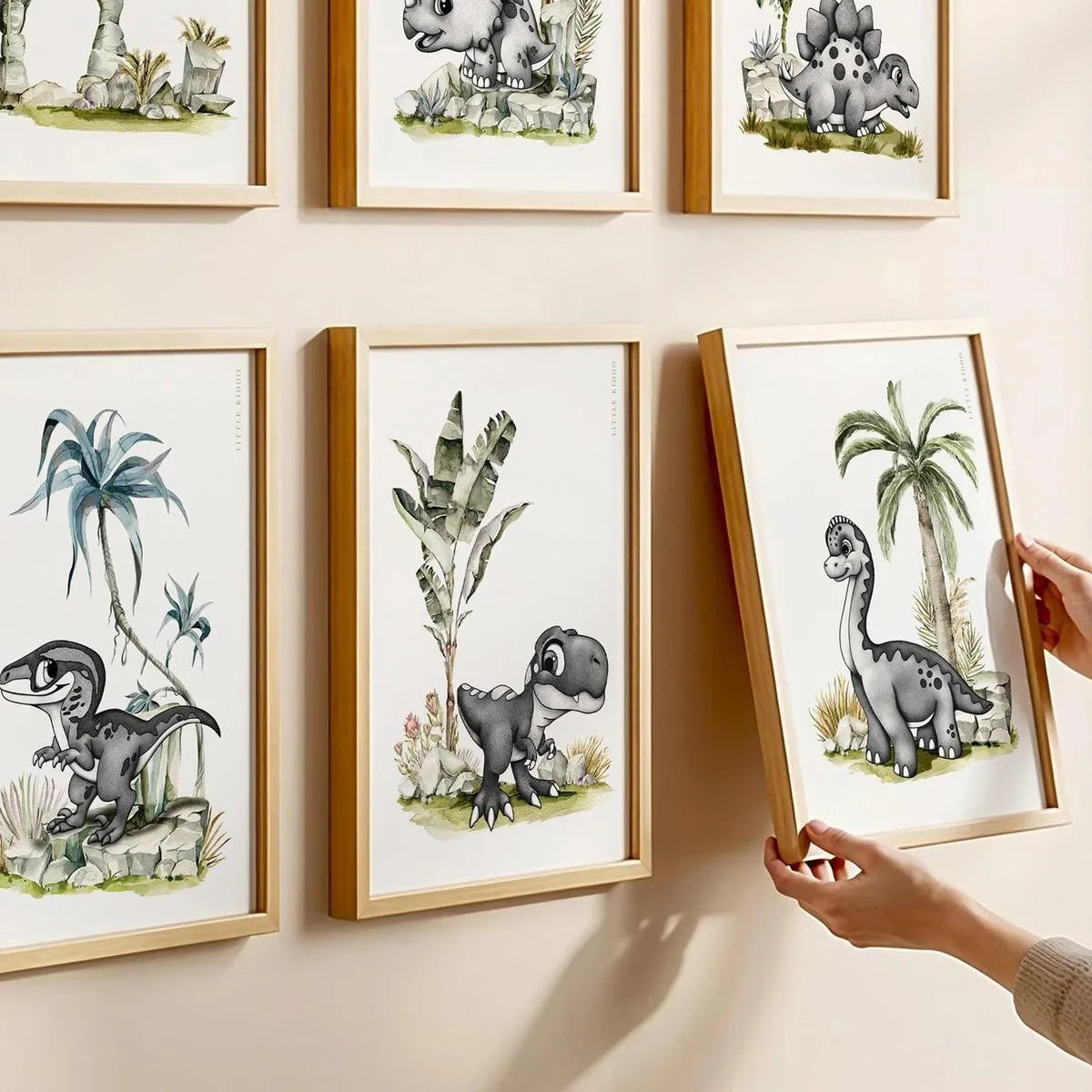 Close-up of hands hanging a baby Brachiosaurus art print as part of a 6-piece dinosaur nursery gallery wall featuring T-Rex and Velociraptor in matching light oak frames.