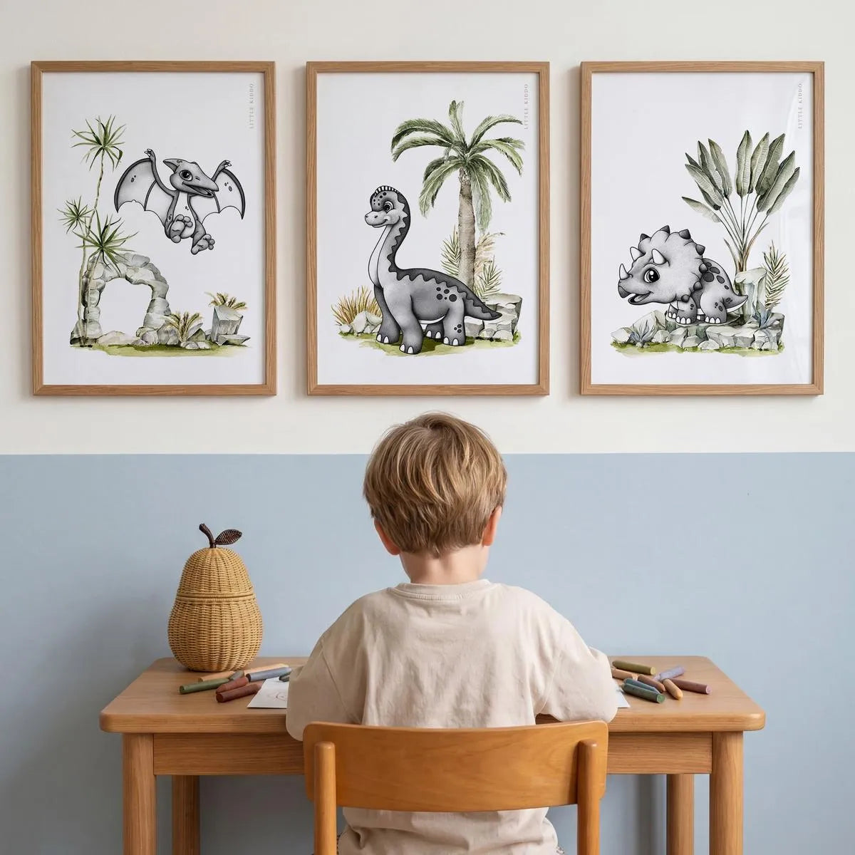 Set of 3 dinosaur nursery prints—Pterodactyl, Brachiosaurus, and Triceratops—in light wood frames, styled above a child's wooden desk in a modern blue and white playroom.