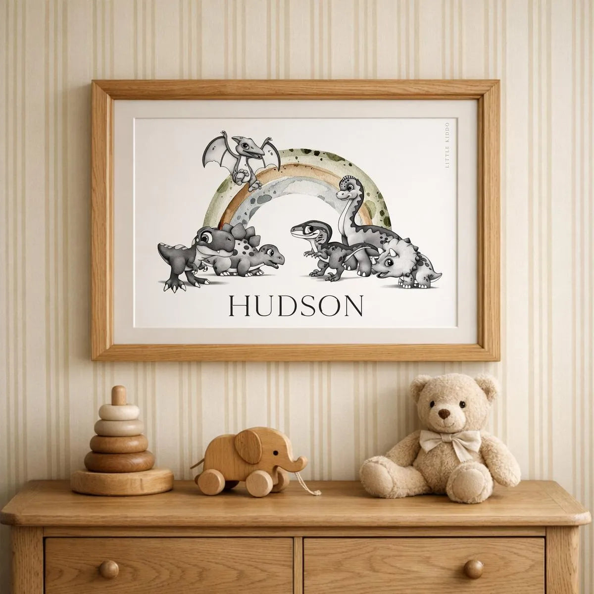 Personalized "Hudson" dinosaur name art in an oak frame, styled on a wooden dresser with a teddy bear and stacking toys in a neutral nursery.