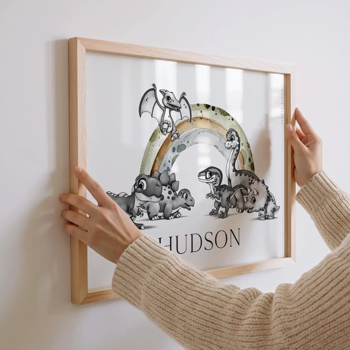 Hands hanging a personalized dinosaur nursery name print for "Hudson" featuring a hand-drawn Pterodactyl, Stegosaurus, and T-Rex under an earthy rainbow.