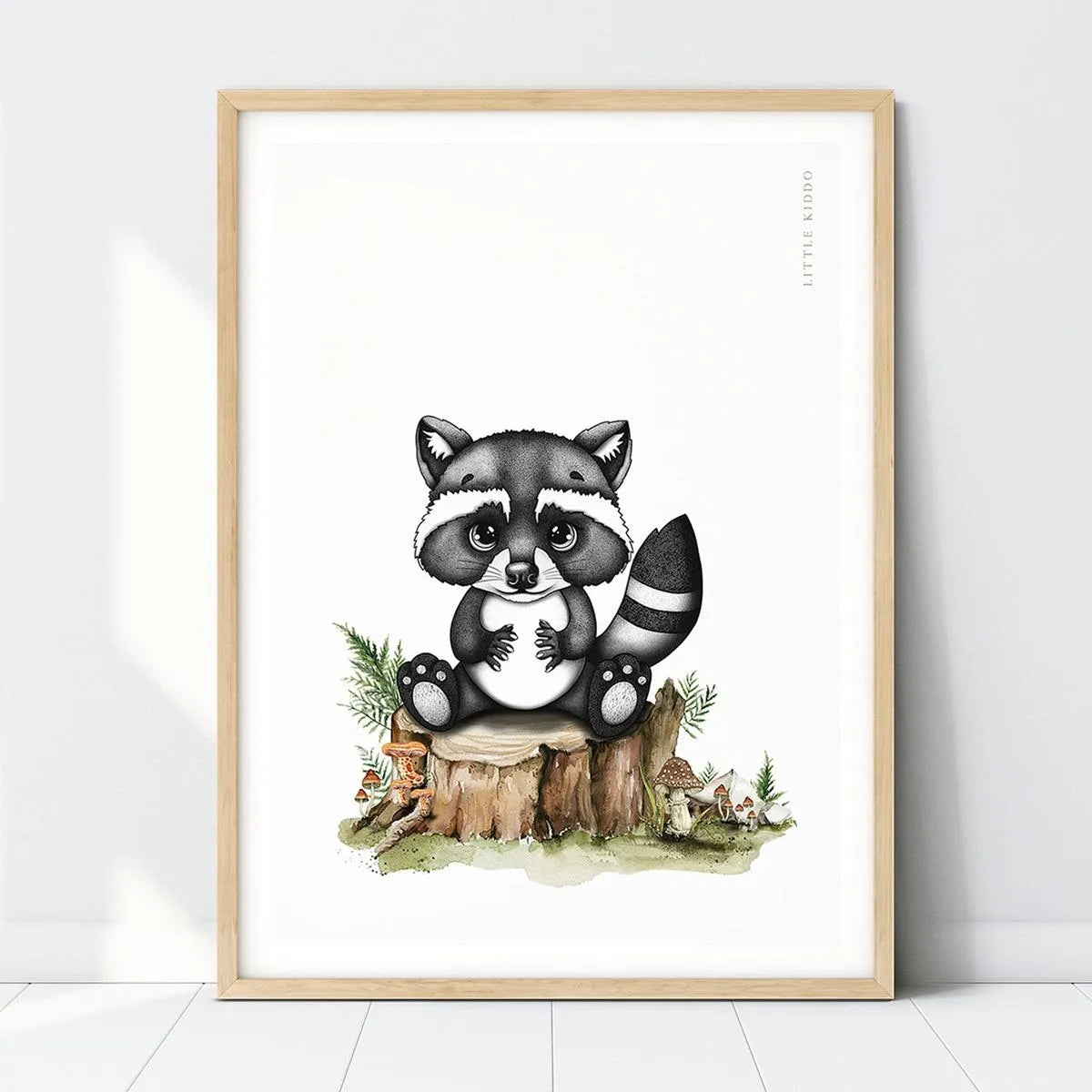 Hand-drawn baby raccoon nursery art print sitting on a tree stump with mushrooms in a light wood frame, woodland forest wall decor.