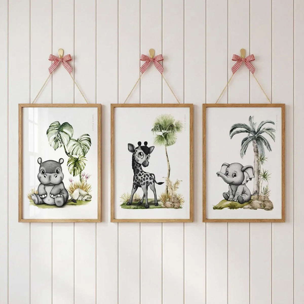 Set of 3 safari nursery prints—hippo, giraffe, and elephant—hanging with pink ribbons on a white wood-paneled wall.