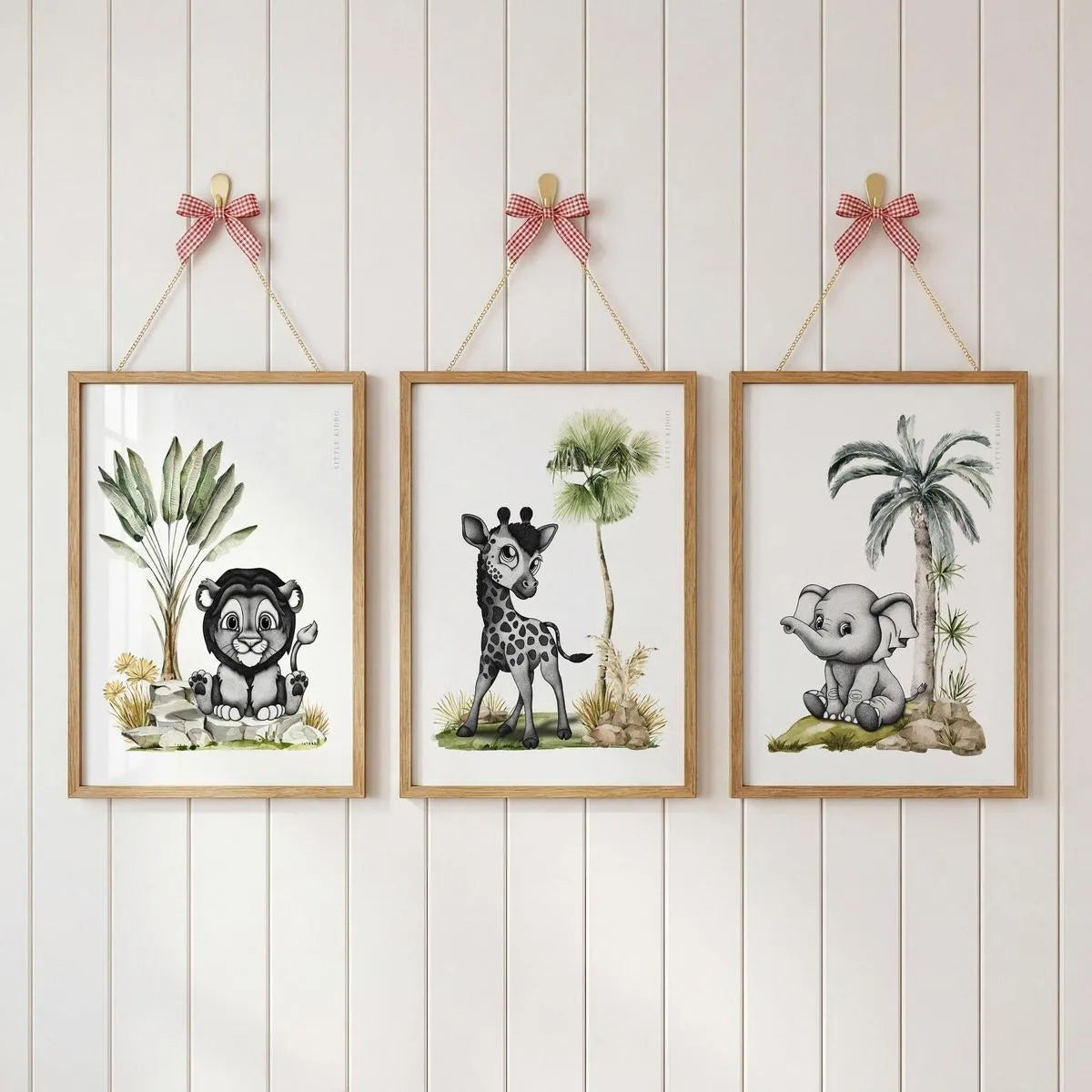 Gallery wall set of 3 safari nursery prints—lion, giraffe, and elephant—hanging with pink ribbons on a white paneled wall.