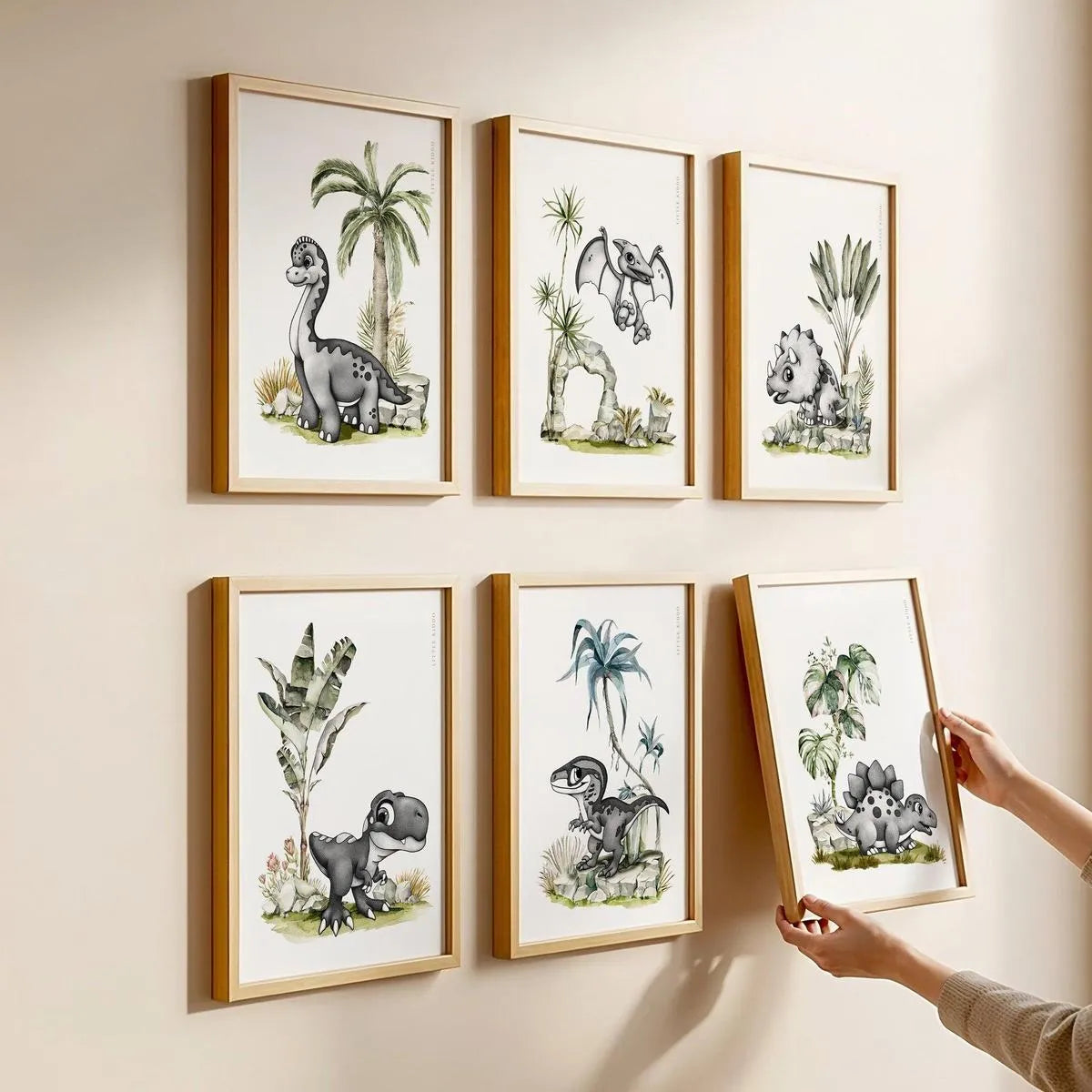 A person hanging a set of 6 dinosaur nursery prints in oak frames, creating a curated Jurassic-themed gallery wall for a playroom.