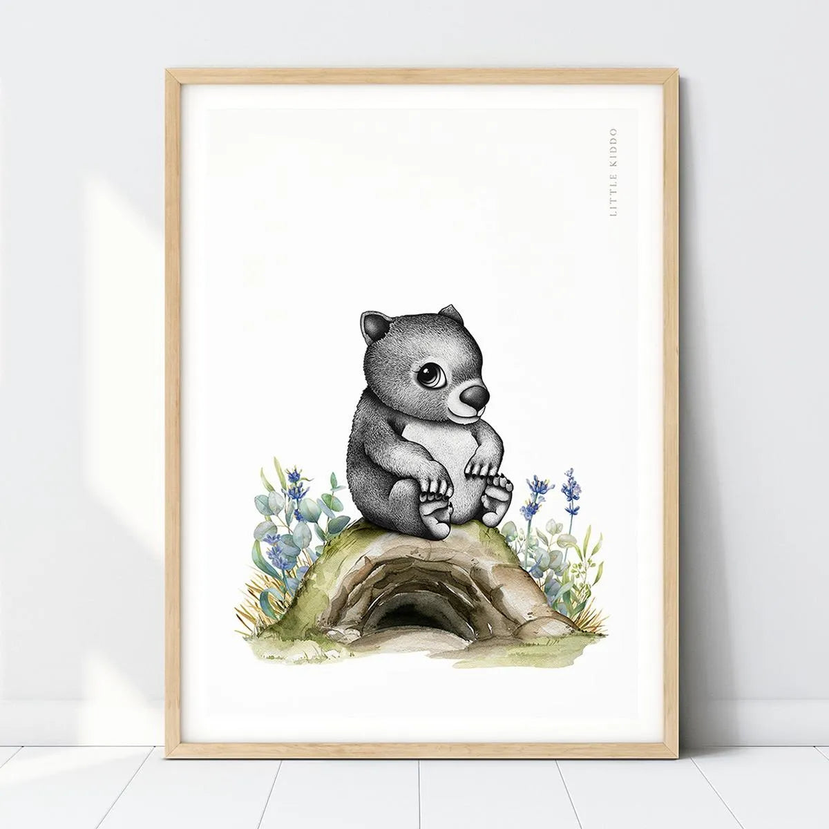 Hand-drawn baby wombat nursery art print sitting on a burrow with blue flowers in a light wood frame, Australian bush animal wall decor.